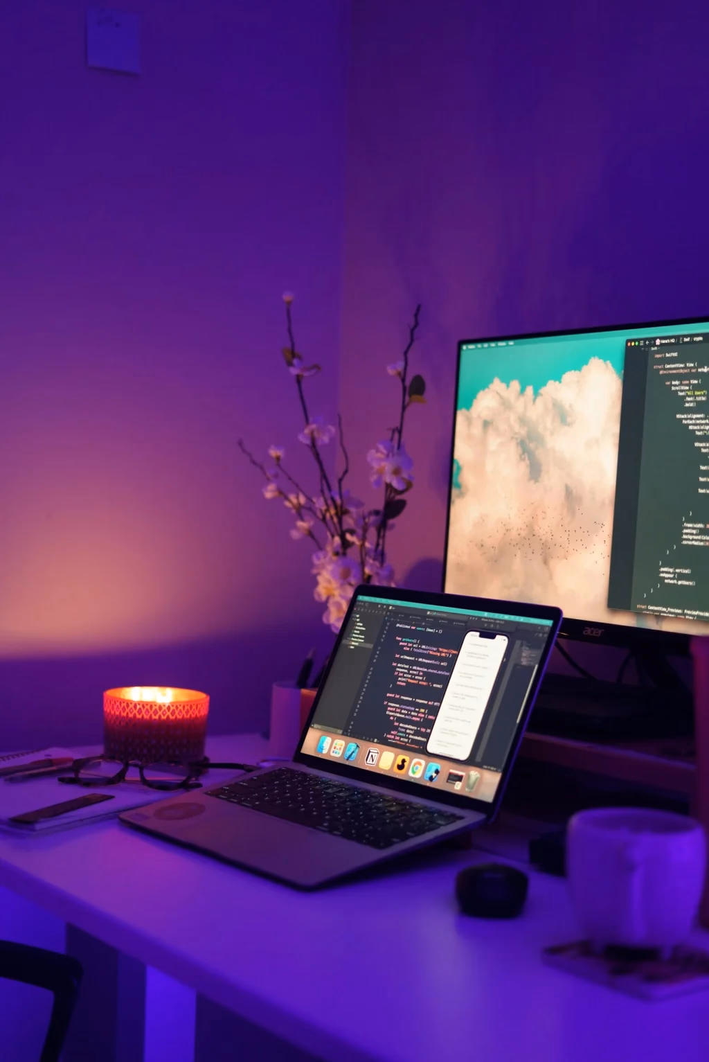 A desk at night lit violet: an external monitor and a laptop both open to code, a candle, and a spray of blossom in a vase.